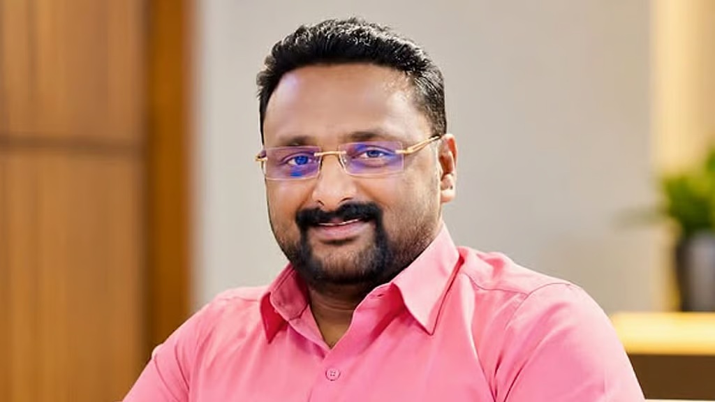 Smiling man with short black hair, beard, and glasses wearing a pink button-down shirt indoors.