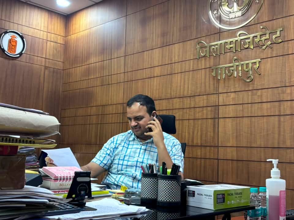 anupam shukla ias | No. 1 Indian Media News Portal Man in a checkered shirt sits at a cluttered desk, talking on a mobile phone while reading a document in a wood-paneled office with Hindi signage on the wall.