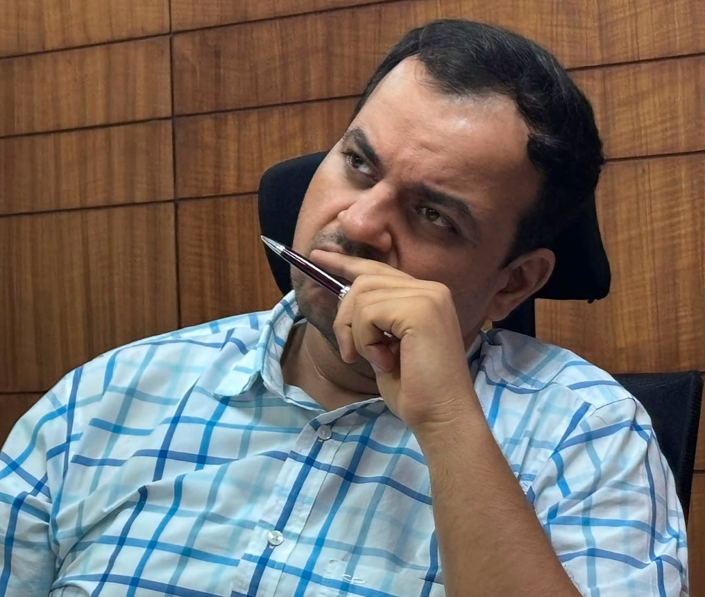 Man in a blue plaid shirt sits in an office chair, pensively resting his hand on his lips with a pen near his mouth.