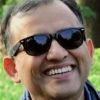 Smiling man wearing dark sunglasses outdoors with greenery in the background.
