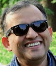 Smiling man wearing dark sunglasses outdoors with greenery in the background.