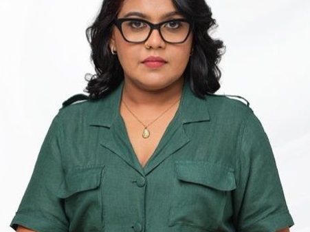Portrait of a woman with dark hair and glasses, wearing a green short-sleeve button-down dress with a belt, posing against a white background.