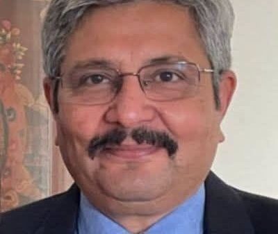 Professional head-and-shoulders portrait of a middle-aged man with gray hair and glasses, wearing a dark suit and blue shirt with a tie, smiling slightly.