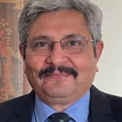 Professional head-and-shoulders portrait of a middle-aged man with gray hair and glasses, wearing a dark suit and blue shirt with a tie, smiling slightly.