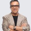 Man with short gray hair and black-r framed glasses wearing a beige textured blazer over a black shirt, arms crossed, smiling.