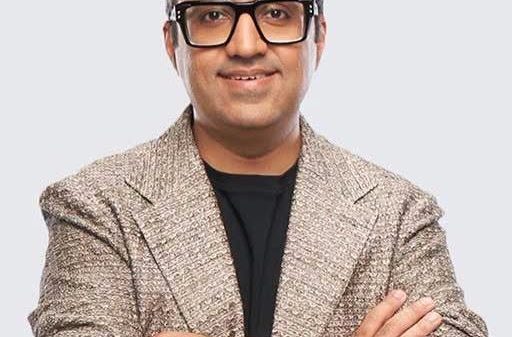 Man with short gray hair and black-r framed glasses wearing a beige textured blazer over a black shirt, arms crossed, smiling.