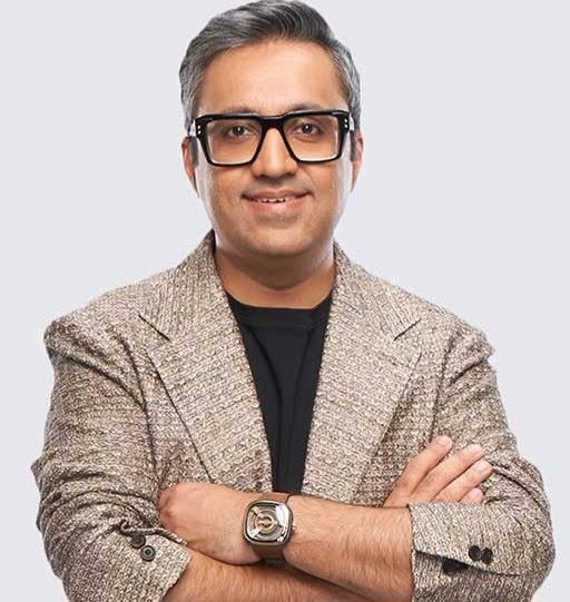 Man with short gray hair and black-r framed glasses wearing a beige textured blazer over a black shirt, arms crossed, smiling.