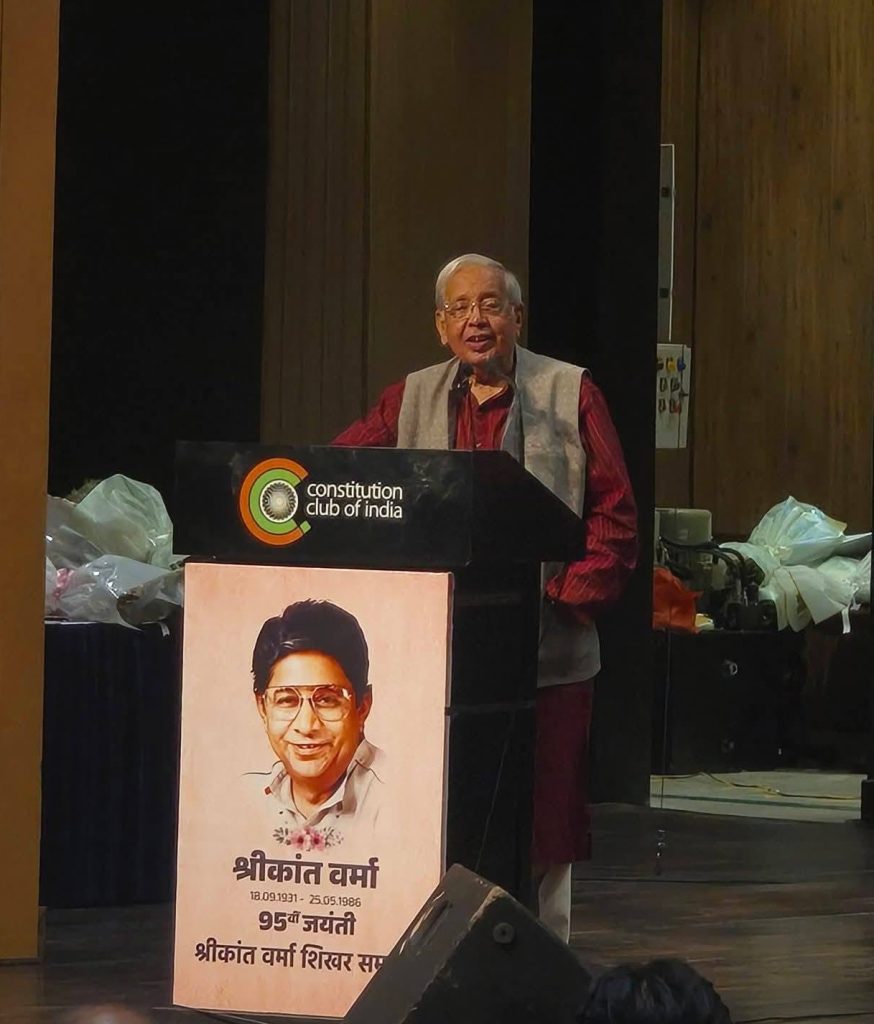 ashok | No. 1 Indian Media News Portal Elderly man speaking at a podium with a Constitution Club of India emblem; a portrait poster with Hindi text stands in front of him.