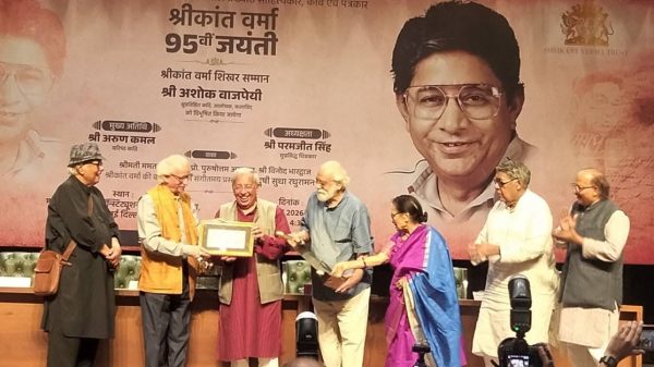Group of seven people on a stage presenting a framed plaque, with a large portrait banner behind them.