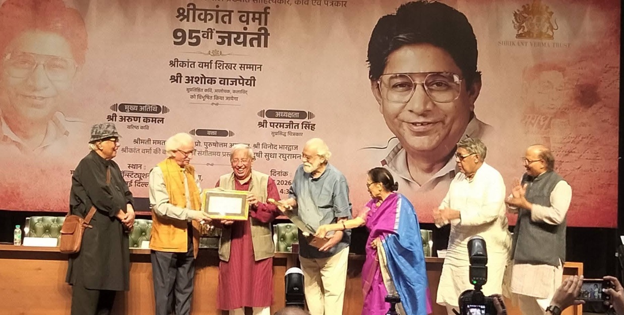 Group of seven people on a stage presenting a framed plaque, with a large portrait banner behind them.