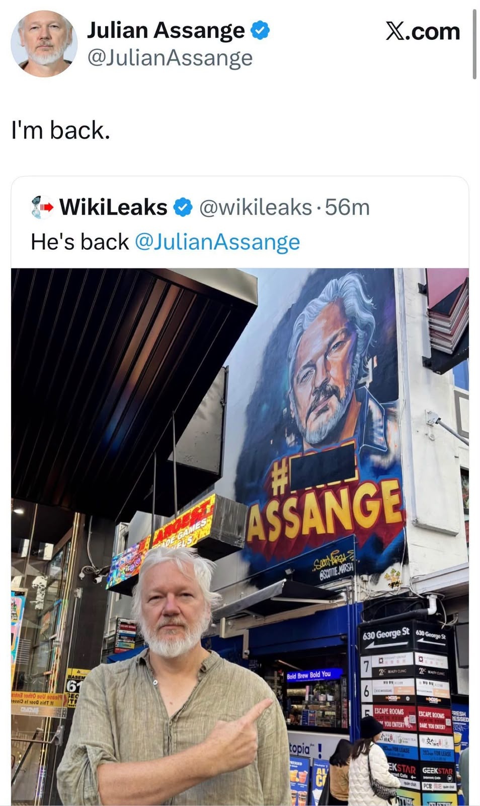 Bearded man in a light shirt stands on a busy city street beside a large mural portrait of a man, with screenshots of tweets about his return above.