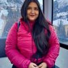Woman in a bright pink puffer jacket sits by a window with snowy mountains in the background, looking at the camera with a slight smile.