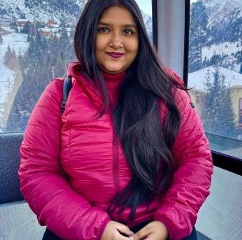 Woman in a bright pink puffer jacket sits by a window with snowy mountains in the background, looking at the camera with a slight smile.