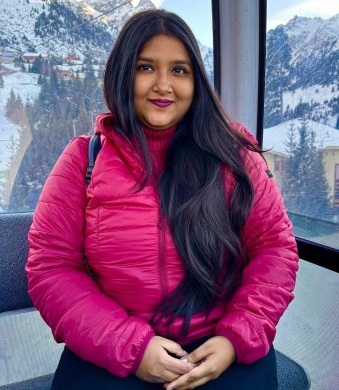 Woman in a bright pink puffer jacket sits by a window with snowy mountains in the background, looking at the camera with a slight smile.