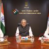 Three Indian election officials sit at a conference table in the Election Commission of India, with flags and a dark backdrop behind them.