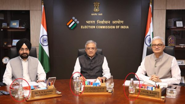 Three Indian election officials sit at a conference table in the Election Commission of India, with flags and a dark backdrop behind them.