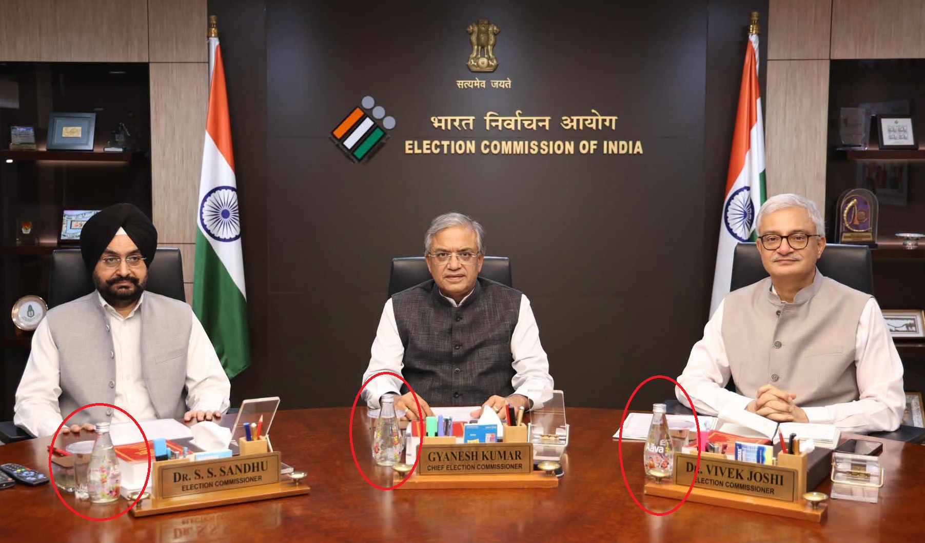 Three Indian election officials sit at a conference table in the Election Commission of India, with flags and a dark backdrop behind them.