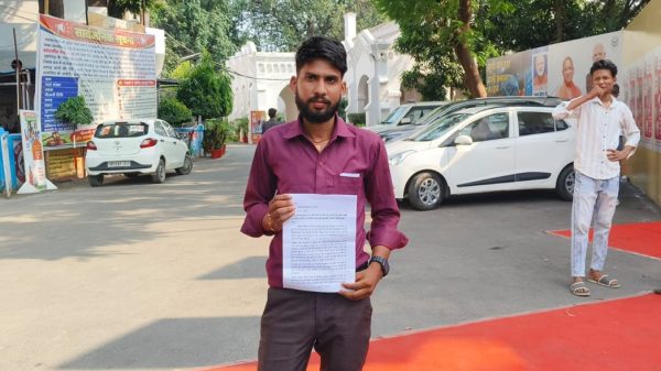 Man in a maroon shirt holding a printed document outdoors, with banners and a white car in the background.