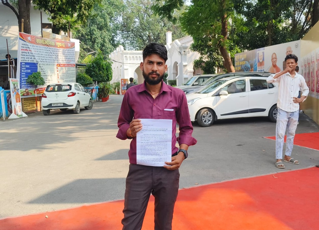 Man in a maroon shirt holding a printed document outdoors, with banners and a white car in the background.