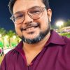 Portrait of a smiling man with glasses at night, wearing a maroon shirt, outdoor lights in the background.