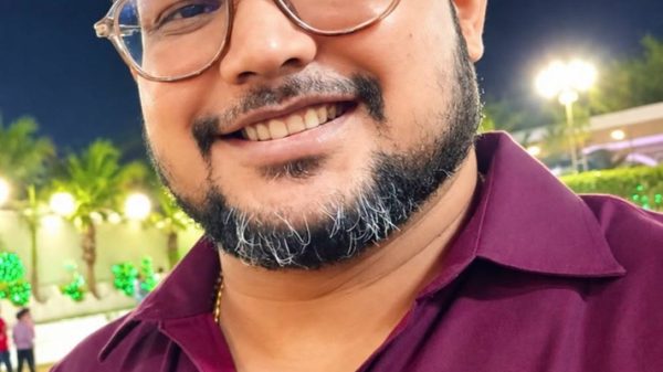 Portrait of a smiling man with glasses at night, wearing a maroon shirt, outdoor lights in the background.