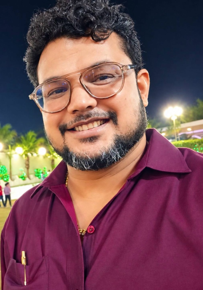 Portrait of a smiling man with glasses at night, wearing a maroon shirt, outdoor lights in the background.