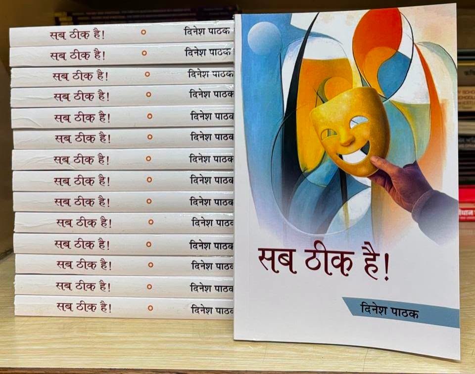 Row of copies of a Hindi book titled 'सब ठीक है!' placed upright beside a colorful cover showing a hand holding a yellow theater mask.