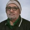 Portrait of an older man wearing glasses, a beige beanie, and a green collared shirt under a dark jacket, with a neutral expression against a pale blue background.