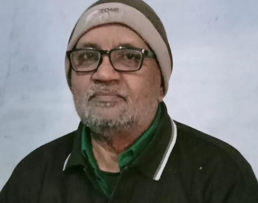 Portrait of an older man wearing glasses, a beige beanie, and a green collared shirt under a dark jacket, with a neutral expression against a pale blue background.