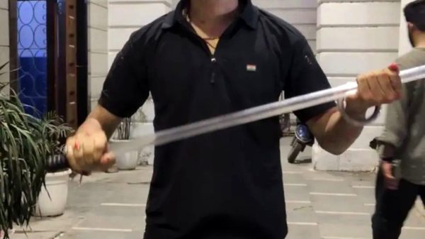 Man in a black polo and sunglasses holds a long metal rod across his chest in an outdoor plaza out front of a white arched building.