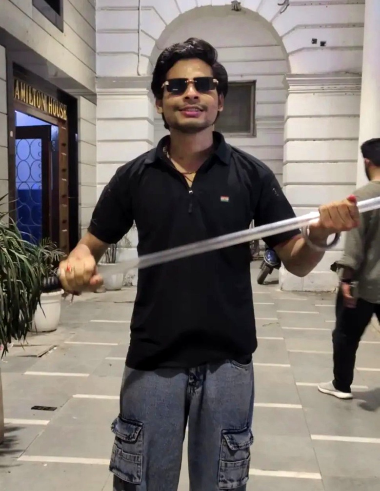 Man in a black polo and sunglasses holds a long metal rod across his chest in an outdoor plaza out front of a white arched building.