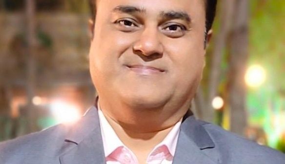 Smiling man in a light gray suit and pink shirt at an outdoor evening event.
