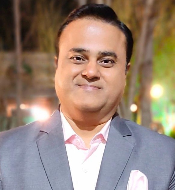 Smiling man in a light gray suit and pink shirt at an outdoor evening event.