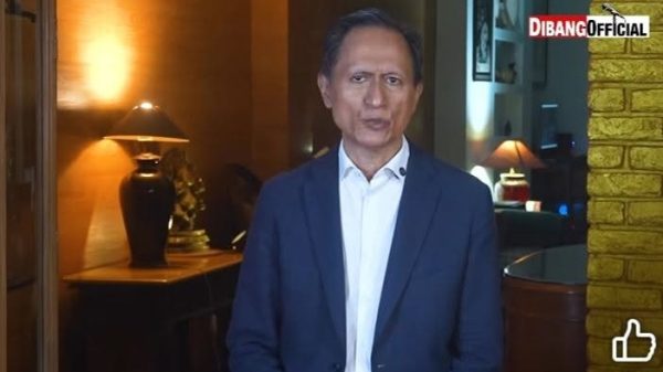 Man in a blue suit and white shirt stands indoors with warm lighting, brick wall on the right, and a watermark reading 'DIBANGOFFICIAL' in the top-right corner.