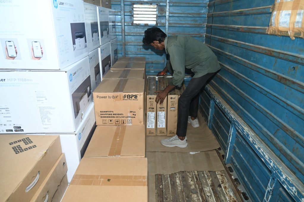 Man in a green shirt bends down to arrange boxes of electronics inside a blue cargo truck.