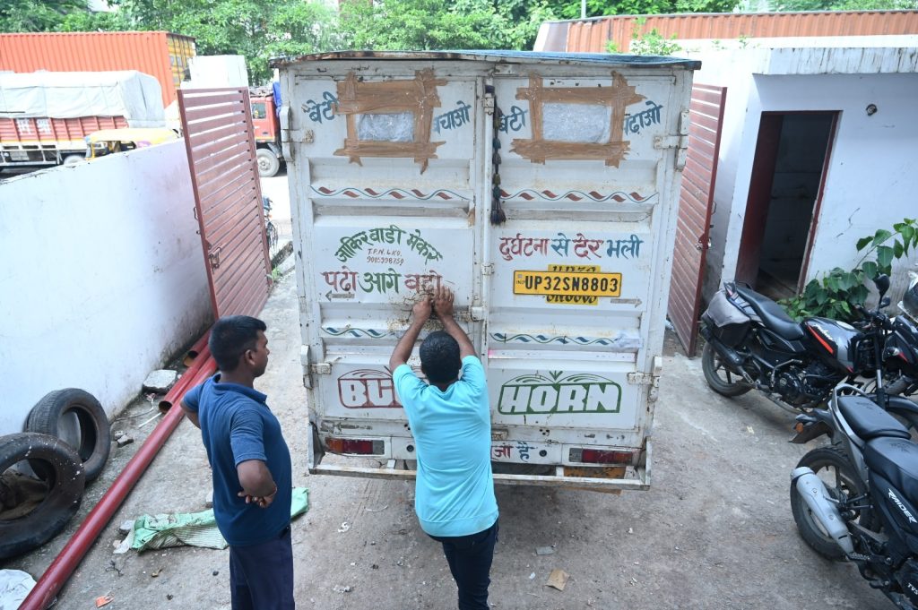 Two men stand at the back of a white truck trailer, tapping a latch as they work, with Hindi text and a yellow license plate visible on the doors, motorcycles nearby.