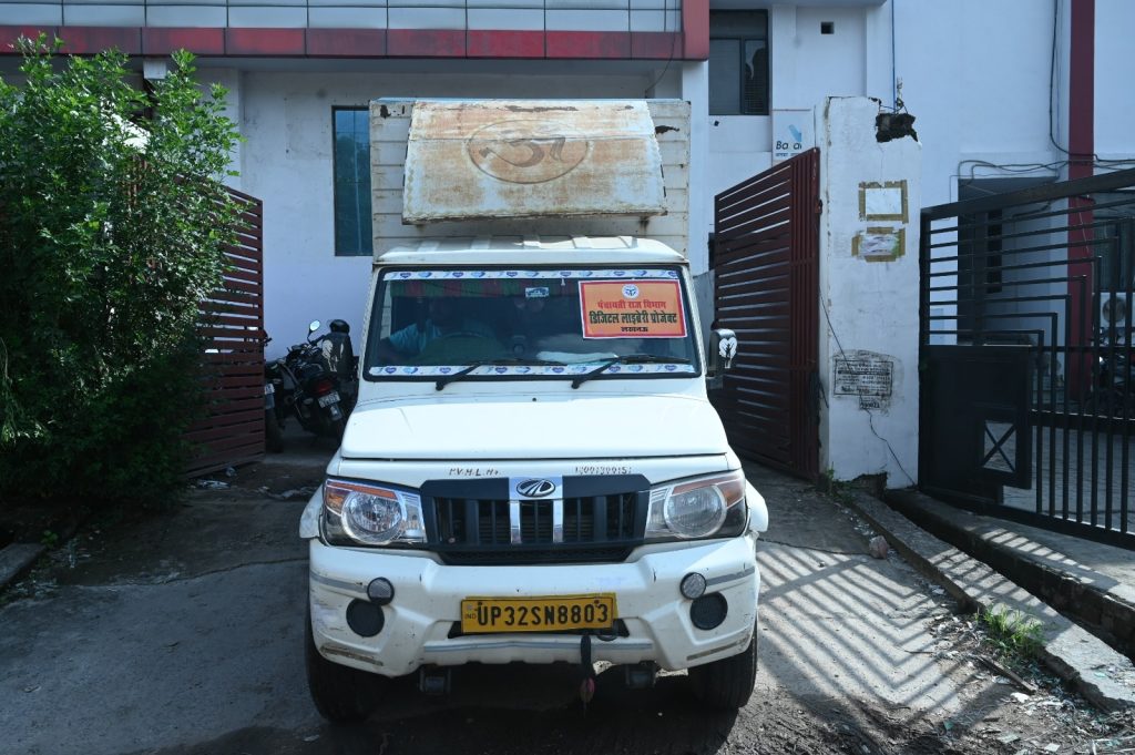 White delivery truck parked in a gated compound, with a large rusty cargo box on top and a yellow Indian license plate UP32SN8803 (UP region). A red sign with Hindi text is visible on the windshield.