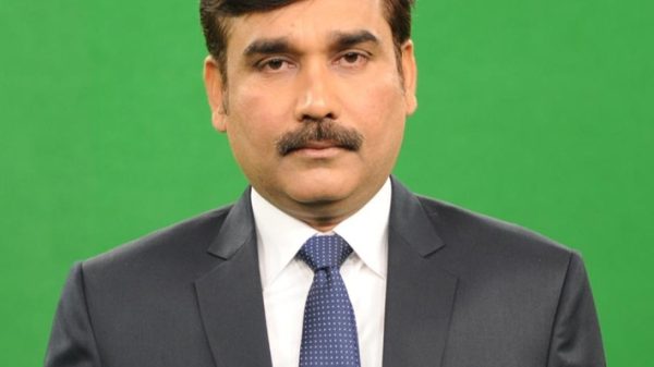 Man in a dark suit with white shirt and blue polka-dot tie, standing against a green backdrop.
