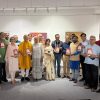 Group of about 20 people in a bright gallery, many in traditional Indian attire, proudly holding red plaques and posing for a group photo.