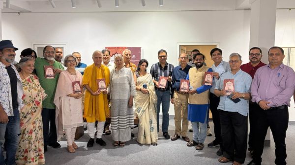 Group of about 20 people in a bright gallery, many in traditional Indian attire, proudly holding red plaques and posing for a group photo.