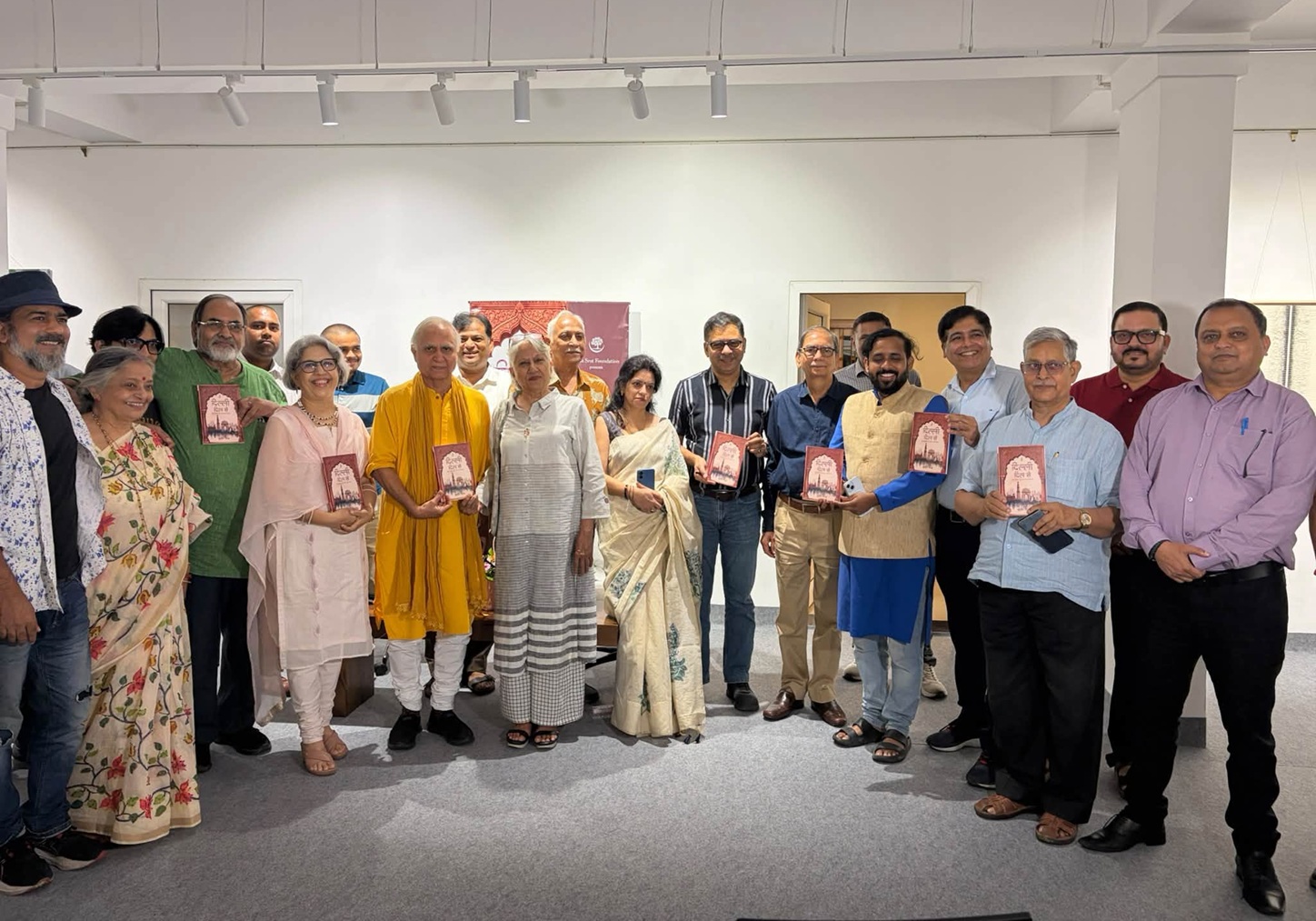 Group of about 20 people in a bright gallery, many in traditional Indian attire, proudly holding red plaques and posing for a group photo.