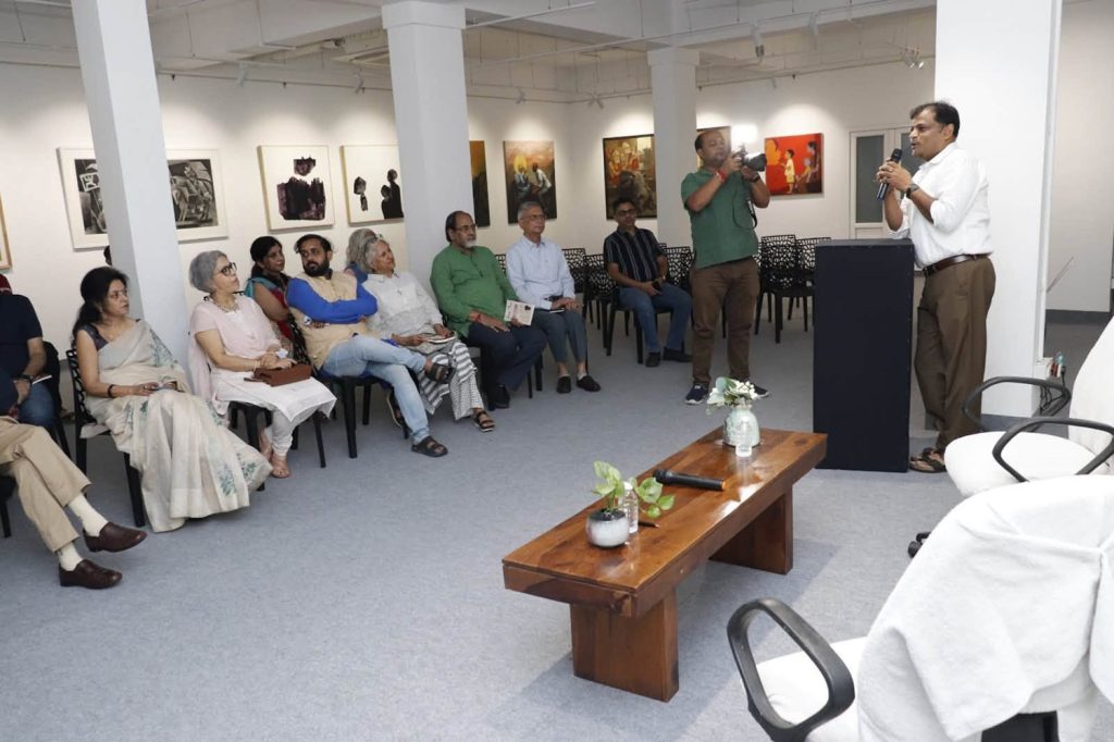 Audience seated in an art gallery listening to a speaker at a podium; a photographer is taking pictures in the background.