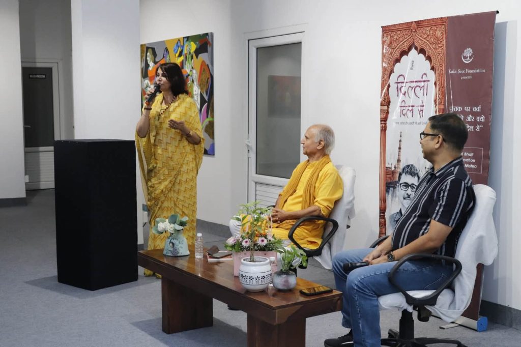 Woman in a yellow sari speaks into a microphone at a podium while two men listen nearby as banners and plants decorate the room.