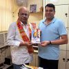Two men in an office exchange a small booklet; one wears a patterned shirt and a colorful scarf, the other in a light blue polo. A fan and posters sit in the background.