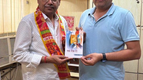 Two men in an office exchange a small booklet; one wears a patterned shirt and a colorful scarf, the other in a light blue polo. A fan and posters sit in the background.