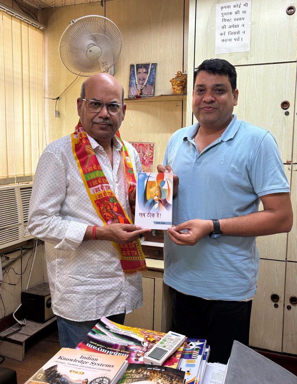 Two men in an office exchange a small booklet; one wears a patterned shirt and a colorful scarf, the other in a light blue polo. A fan and posters sit in the background.