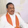 Man in a white shirt with an orange striped scarf, seated on a beige sofa, and wearing a tilak on his forehead.