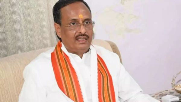 Man in a white shirt with an orange striped scarf, seated on a beige sofa, and wearing a tilak on his forehead.
