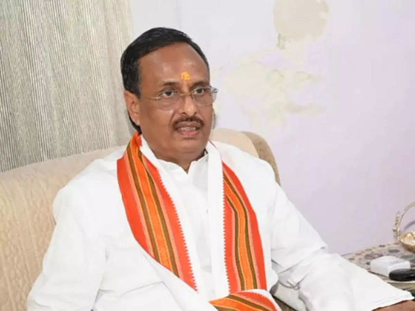 Man in a white shirt with an orange striped scarf, seated on a beige sofa, and wearing a tilak on his forehead.