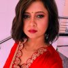 Portrait of a woman with short dark hair wearing a red traditional top with gold floral embroidery and a green bindi looking at the camera.